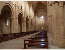 Interno di una chiesa in pietra con navata centrale e file di panche in legno, delimitata da arcate su colonne massicce e volte a crociera. In fondo si vede un rosone circolare e finestre strette che illuminano l’abside.
