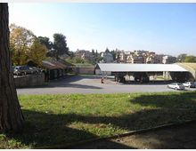 Area asfaltata con pensiline basse in metallo, simile a un parcheggio o deposito, vista da un prato in pendenza con alberi ai lati. Sullo sfondo si vede un quartiere collinare con edifici residenziali e vegetazione sotto un cielo sereno.