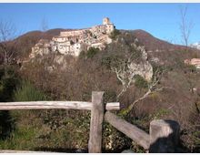 Borgo storico arroccato su una collina, con case in pietra addossate tra loro e una torre in cima. In primo piano si vede una staccionata in legno e vegetazione invernale, con colline boscose sullo sfondo sotto un cielo sereno.
