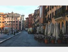 Strada lastricata in un centro storico con alti palazzi dai colori pastello e finestre con persiane. Sul lato destro si vedono tavolini all’aperto con sedie e grandi ombrelloni chiusi, affacciati su una piazza.