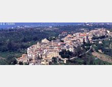 Veduta panoramica aerea di un borgo collinare con case in pietra e tetti in laterizio addossate tra loro, circondato da aree boscate e campi. Sullo sfondo si estende il paesaggio rurale con colline e vegetazione.