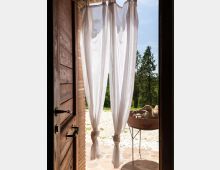 Porta in legno aperta su un patio in pietra, con tende bianche leggere che pendono e filtrano la luce. All’esterno si vede un cortile ghiaioso con un braciere su supporto metallico e alberi sullo sfondo sotto un cielo sereno.