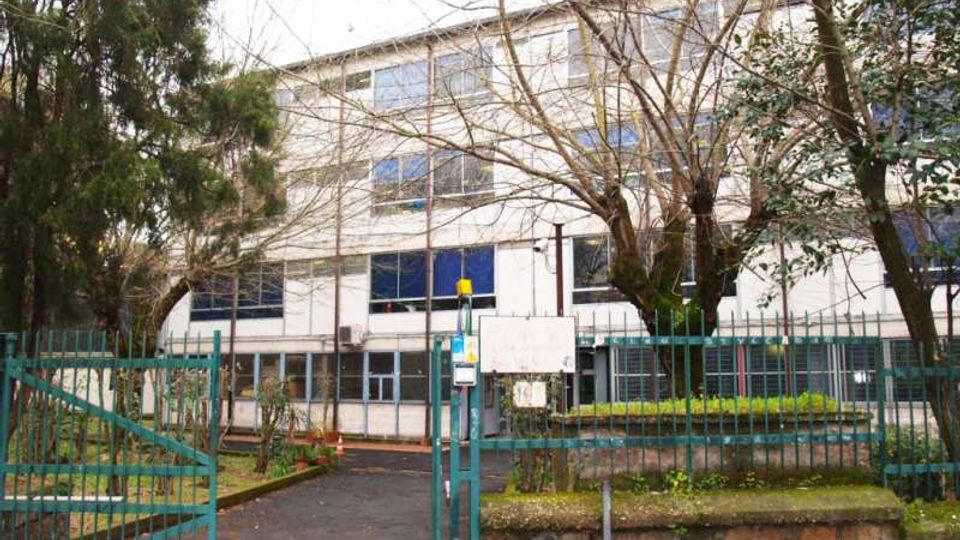 Edificio scolastico a più piani con facciata chiara e ampie finestre, visto oltre una recinzione metallica verde con cancello d’ingresso. Davanti si trovano un vialetto asfaltato, aiuole rialzate in muratura e alberi che incorniciano l’area esterna.