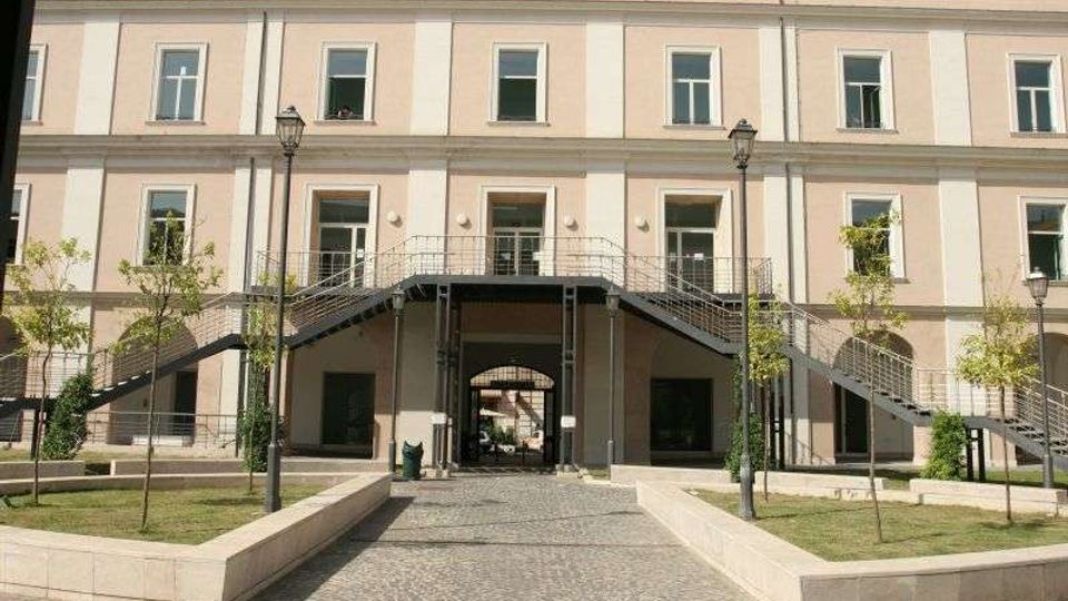 Facciata di un edificio a tre piani color crema con finestre regolari e un grande arco centrale d’ingresso. Davanti si vede un cortile con vialetto in pietra, aiuole con giovani alberi e una passerella sopraelevata con scale metalliche simmetriche sui due lati.