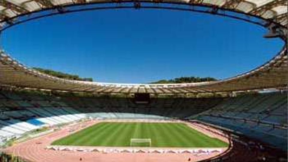 Veduta interna di un grande stadio con copertura ellittica e gradinate che circondano un campo da calcio in erba, con pista di atletica rossa tutt’intorno. Il cielo azzurro è visibile attraverso l’ampia apertura centrale del tetto.