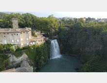 Veduta aerea di un borgo con edifici in pietra e una torre medievale affacciati su una gola boscosa. Al centro, una cascata scende in un bacino d’acqua verde circondato da pareti rocciose e vegetazione fitta.