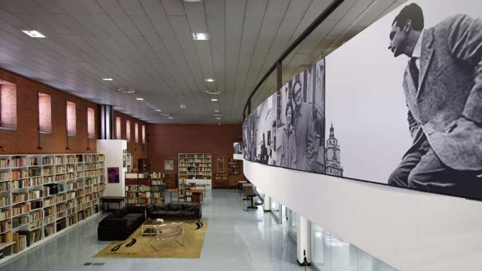 Interno di una biblioteca moderna con alte scaffalature di libri lungo pareti in mattoni e ampie finestre verticali. A destra corre un ballatoio curvo con grandi pannelli fotografici in bianco e nero, mentre al centro si trova un’area lettura con divani e tavolini su un tappeto.