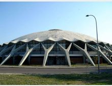 Vista frontale di un grande palazzetto dello sport con ampia copertura a cupola e strutture portanti esterne in cemento a forma di archi inclinati. Davanti si vede una strada con marciapiede e lampione, sotto un cielo sereno.