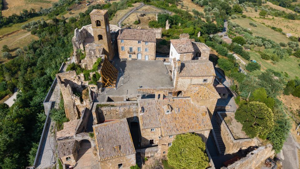 Veduta aerea di un borgo fortificato in pietra con mura perimetrali, edifici dai tetti in coppi e una torre campanaria, con alcune strutture in rovina. Il complesso è circondato da colline rurali con campi coltivati, uliveti e aree boscate.