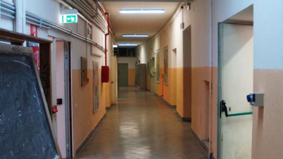 Corridoio interno lungo in un edificio scolastico o industriale, con pavimento piastrellato grigio, pareti bicolore e illuminazione a neon. Sono visibili tubazioni a vista sul soffitto, porte laterali e un cartello di uscita di emergenza illuminato.