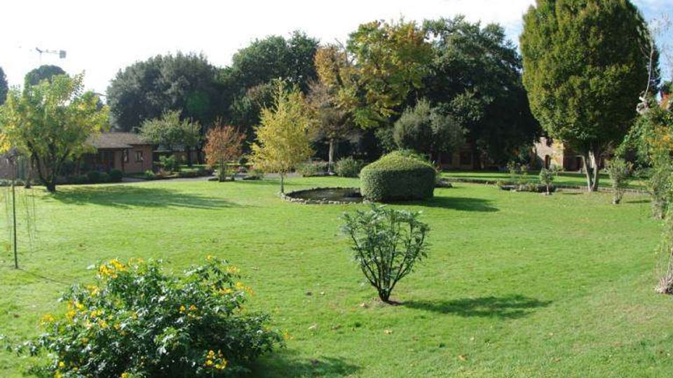 Ampio giardino con prato verde curato, cespugli e piccoli alberi sparsi, con una siepe arrotondata vicino a una piccola area d’acqua. Sullo sfondo si vedono alberi alti e alcune strutture basse immerse nel verde.