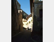 Vicolo stretto in salita con pavimentazione in pietra, fiancheggiato da alte mura in pietra e da edifici storici con intonaco chiaro e tetti in tegole. Sullo sfondo si vede una grande struttura fortificata in pietra sotto un cielo sereno.