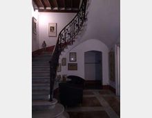 Interno di un edificio con ampia scala in pietra che sale a sinistra e ringhiera in ferro battuto decorata. Il pianerottolo e il corridoio sottostante hanno pareti bianche con quadri incorniciati, arco di passaggio e pavimento in piastrelle.