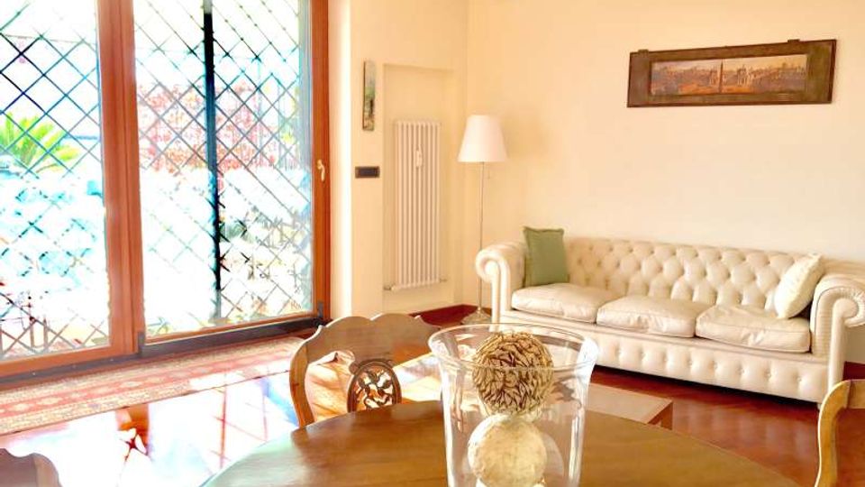 Soggiorno luminoso con pavimento in legno lucido, grande porta finestra scorrevole con grata esterna e vista sul giardino. Nella stanza ci sono un divano chiaro stile Chesterfield, un tavolo rotondo in legno con sedie e una lampada da terra vicino alla parete con quadro.