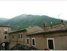 Case in muratura con facciate color ocra e tetti in coppi, con finestre a persiane scure e un piccolo passaggio ad arco. Sullo sfondo si vedono montagne verdi boscose sotto un cielo chiaro.