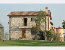 Casale di campagna in pietra a due piani con tetto in coppi, balcone con ringhiera e scala esterna ad arco; davanti un prato curato con alberelli e vasi. Sullo sfondo si vedono elementi di un giardino e una struttura cilindrica color rosa.