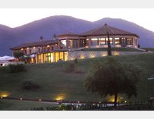 Edificio tipo resort o agriturismo su una collina erbosa, con tetti in coppi e grandi vetrate illuminate. Sullo sfondo si vedono montagne al crepuscolo e, in primo piano, alberi e un vialetto con luci a terra.