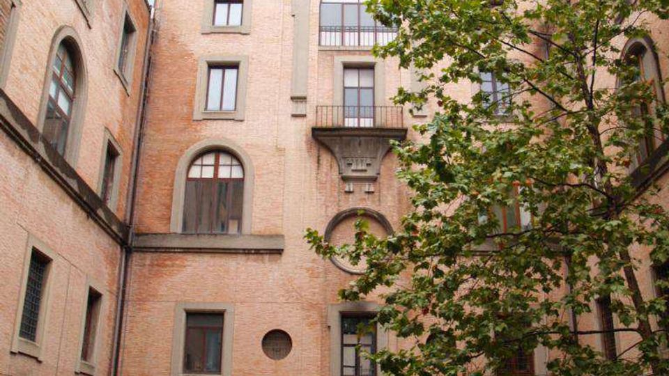 Cortile interno con edificio in mattoni a più piani, con finestre ad arco e un grande arco decorativo centrale sopra un balconcino. Un albero occupa il lato destro e l’ingresso al piano terra è coperto da una tettoia, con area pavimentata e auto parcheggiate.