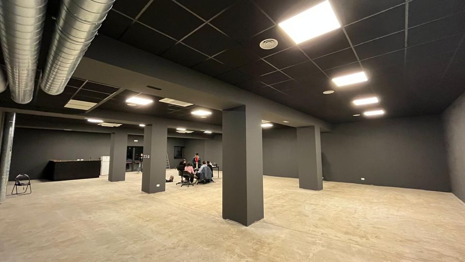 Ampio spazio interno tipo ufficio o sala polifunzionale con pareti e colonne grigio scuro, pavimento in cemento e soffitto nero a griglia con luci a pannello. Sono visibili condotti di aerazione metallici a vista e un lungo bancone sul lato sinistro.