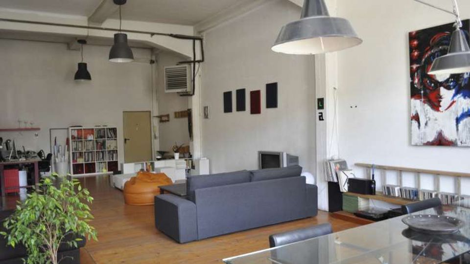 Ampio loft open space con pavimento in legno, soffitto alto con travi e grandi lampade industriali a sospensione. Zona soggiorno con divano grigio e pouf, tavolo in vetro e scaffali con libri e dischi lungo le pareti bianche decorate con quadri.