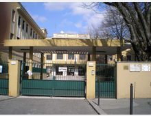 Ingresso di un edificio pubblico con cancello metallico verde e pilastri gialli, coperto da una tettoia orizzontale che conduce al cortile interno. Sullo sfondo si vede la facciata dell’edificio con finestre e bandiere, affiancata da alberi spogli lungo il perimetro.