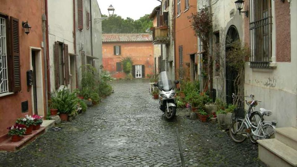 Vicolo acciottolato in un borgo storico, fiancheggiato da case intonacate con persiane in legno, lampioni a parete e numerosi vasi di piante lungo i muri. In primo piano si vedono una moto e una bicicletta parcheggiate, con una piccola piazzetta e un edificio sullo sfondo.