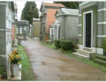 Viale di un cimitero con strada sterrata tra tombe e cappelle funerarie in muratura e pietra, alcune con colonne e porte scure. Ai lati si vedono piccoli cespugli, fiori in vaso e alti alberi sullo sfondo.