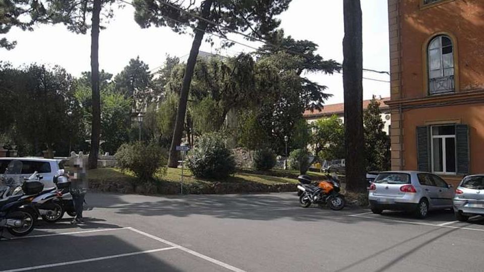 Parcheggio e incrocio stradale davanti a un’area verde con alberi alti e cespugli. Sul lato destro si vede la facciata di un edificio in intonaco arancione con finestre, e lungo la strada sono presenti auto e motociclette parcheggiate.