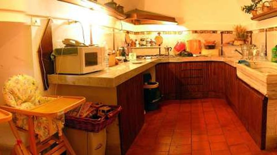 Cucina rustica in mansarda con soffitto spiovente, pavimento in cotto e mobili in legno scuro disposti a L. Piano di lavoro in pietra con lavello e mensole illuminate, piccoli elettrodomestici e due seggioloni con vassoi vicino a una finestra bassa.