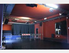 Interno di una piccola sala eventi con pareti rosso scuro e pavimento in piastrelle nere, con palco sul fondo chiuso da tende nere. Al soffitto ci sono tralicci con luci e altoparlanti, e davanti al palco sono presenti transenne metalliche.