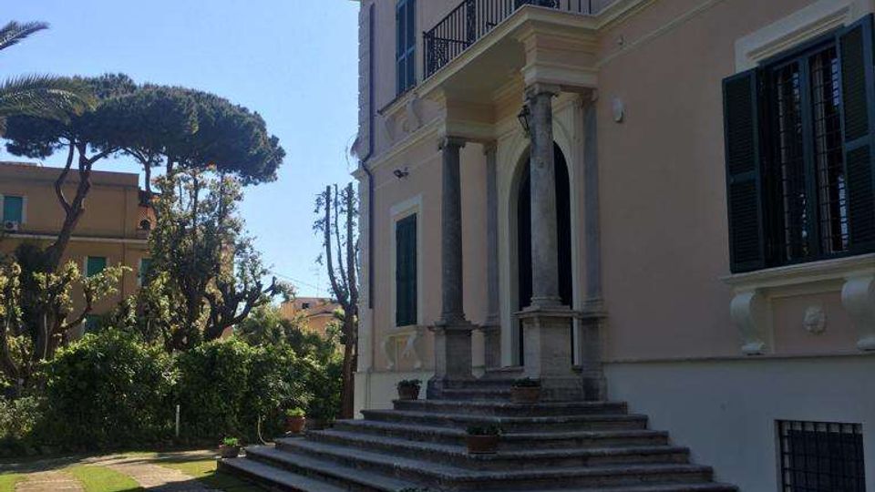 Facciata laterale di una villa storica color rosa con persiane scure, portico d’ingresso con colonne e una scalinata in pietra che conduce a una porta ad arco. Davanti si vede un vialetto lastricato tra strisce di prato e un giardino con alberi, con altri edifici sullo sfondo.