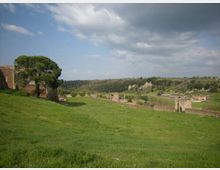 Ampio prato collinare verde con un grande albero in primo piano e resti di mura in pietra con torri e un arco sul lato destro. Sullo sfondo si vedono campi e colline boscose sotto un cielo parzialmente nuvoloso.