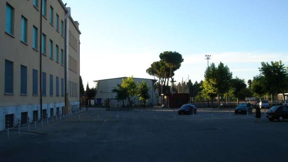 Ampio piazzale asfaltato con alcuni parcheggi e pochi veicoli, affiancato a sinistra da un edificio a più piani con file di finestre. Sullo sfondo si vedono alberi, una struttura bassa tipo capannone e un’area recintata sotto un cielo sereno.