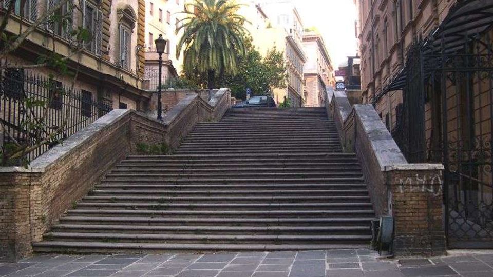Ampia scalinata in pietra con parapetti laterali in mattoni che sale tra edifici storici, con lampioni e cancellate in ferro ai lati. In cima si vedono una palma e alberi che incorniciano la strada sovrastante.