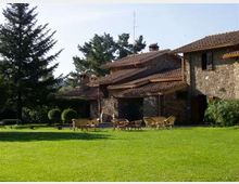 Edificio rurale in pietra con tetti in tegole rosse e portico, affacciato su un ampio prato verde con sedie e tavolini da giardino. Alberi alti e cespugli delimitano l’area esterna sullo sfondo.