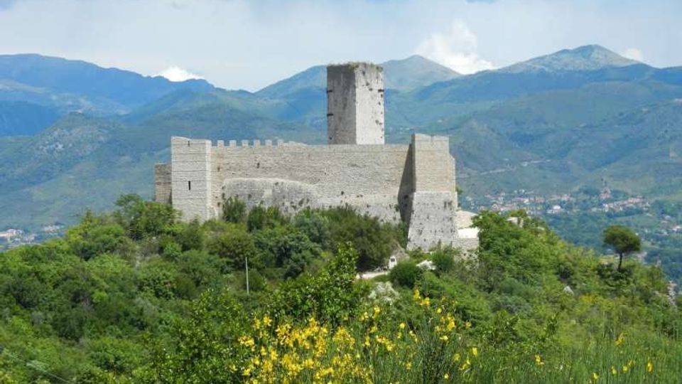 Castello in pietra con mura merlate e torre centrale su una collina, circondato da vegetazione e cespugli fioriti in primo piano. Sullo sfondo si vedono una valle e una catena montuosa sotto un cielo parzialmente nuvoloso.