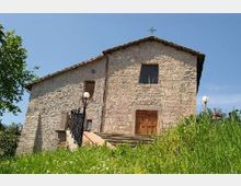 Edificio storico in pietra, simile a una piccola chiesa, con tetto a falde, porta in legno e una croce sulla sommità della facciata. L’ingresso è raggiungibile tramite una scalinata con ringhiera, circondato da erba alta e alberi sotto un cielo sereno.