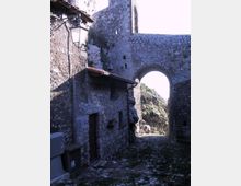 Vicolo stretto in pietra con pavimentazione acciottolata, fiancheggiato da edifici in muratura grezza con piccole finestre e una lanterna a parete. In fondo si apre un grande arco in pietra, che incornicia rocce e vegetazione su un pendio esterno.