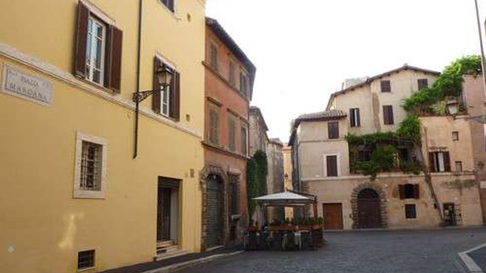 Piazzetta lastricata con edifici storici a più piani dalle facciate color ocra e rosa, con finestre a persiana e portoni ad arco. Al centro c’è un’area con tavoli e ombrelloni, e sullo sfondo si vedono edifici con balconi e vegetazione rampicante.