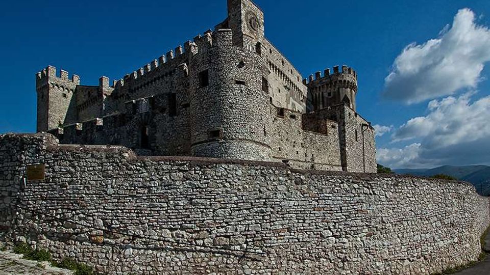 Castello medievale in pietra con merlature e torri, visto dal basso dietro un’alta cinta muraria in pietra. Sullo sfondo cielo azzurro con nuvole e colline lontane.