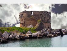 Rovine di una torre in pietra su un piccolo promontorio erboso, circondata da massi lungo la riva. In primo piano si vede l’acqua calma e sullo sfondo un cielo nuvoloso.