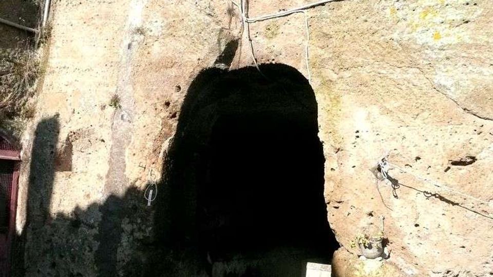 Ingresso a un tunnel scavato nella roccia, con apertura ad arco in una parete di pietra naturale. Sopra l’entrata sono visibili cavi e un faro, con una piccola strada in cemento che conduce all’interno buio.
