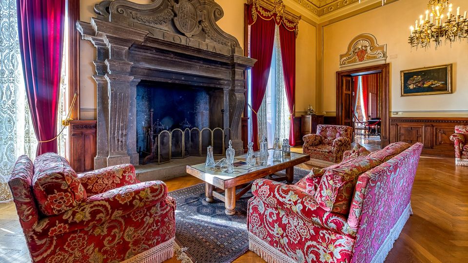 Salone storico in stile classico con grande camino monumentale in pietra scolpita, soffitto decorato e lampadari. Arredi in tessuto rosso a motivi floreali attorno a un tavolino, pavimento in parquet, alte finestre con tende rosse e boiserie in legno lungo le pareti.