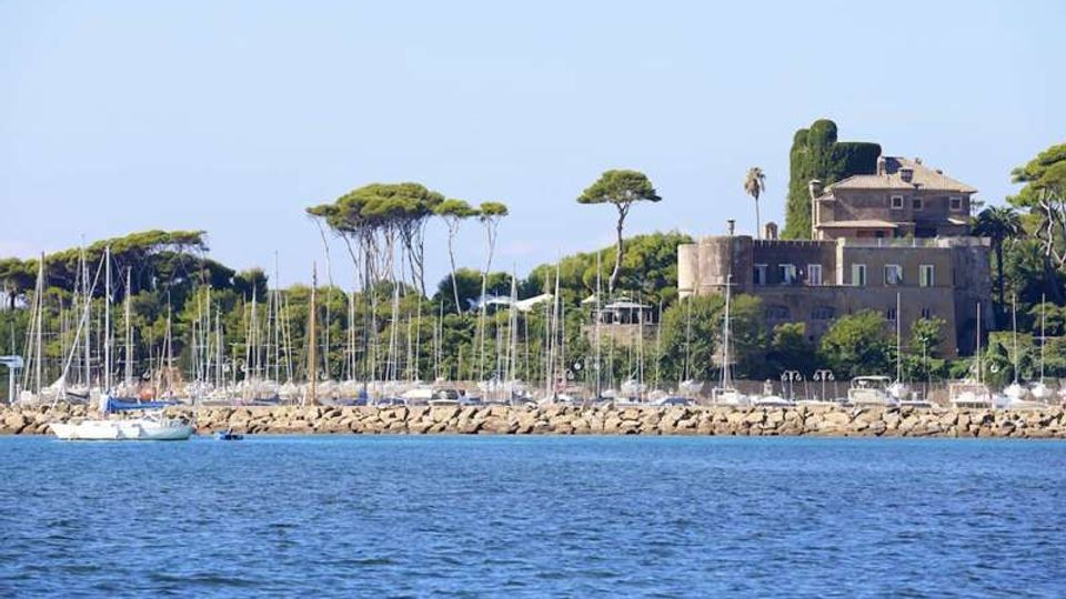 Veduta di un porto turistico con numerose barche a vela ormeggiate dietro una diga frangiflutti in pietra, con il mare in primo piano. Sullo sfondo si vede una grande villa in muratura tra pini marittimi e vegetazione costiera.