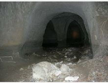 Interno di una grotta o galleria sotterranea con pareti di roccia grigia e volte arrotondate sostenute da colonne naturali. Il pavimento è irregolare, coperto di terra e detriti con alcune pietre e massi in primo piano, e sullo sfondo si intravedono aperture più scure.
