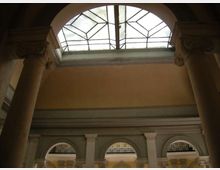Interno di un edificio storico con grandi colonne e arcate inferiori, illuminato da un lucernario a volta con vetrata geometrica. Pareti intonacate e cornici decorative incorniciano l’atrio a doppia altezza.