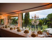 Sala ricevimenti con ampia vetrata panoramica affacciata su un giardino con cipressi e colline sullo sfondo. Lungo tavolo apparecchiato con tovaglia bianca, composizioni floreali, bicchieri e candelabri in vetro trasparente.