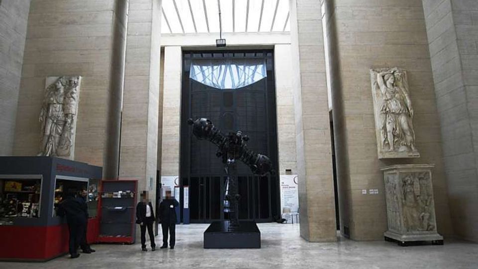 Ampio atrio museale con pareti in pietra chiara e soffitto alto con lucernario, dominato da un grande portale scuro sul fondo. Al centro si trova una scultura metallica su basamento, affiancata da grandi rilievi e statue in pietra lungo le pareti.