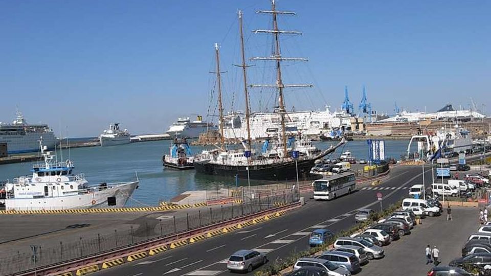 Veduta di un porto con navi ormeggiate, tra cui un grande veliero a più alberi, e altre imbarcazioni lungo la banchina. In primo piano si vede una strada costiera con corsie e parcheggi pieni di auto, con infrastrutture portuali e gru sullo sfondo sotto cielo sereno.