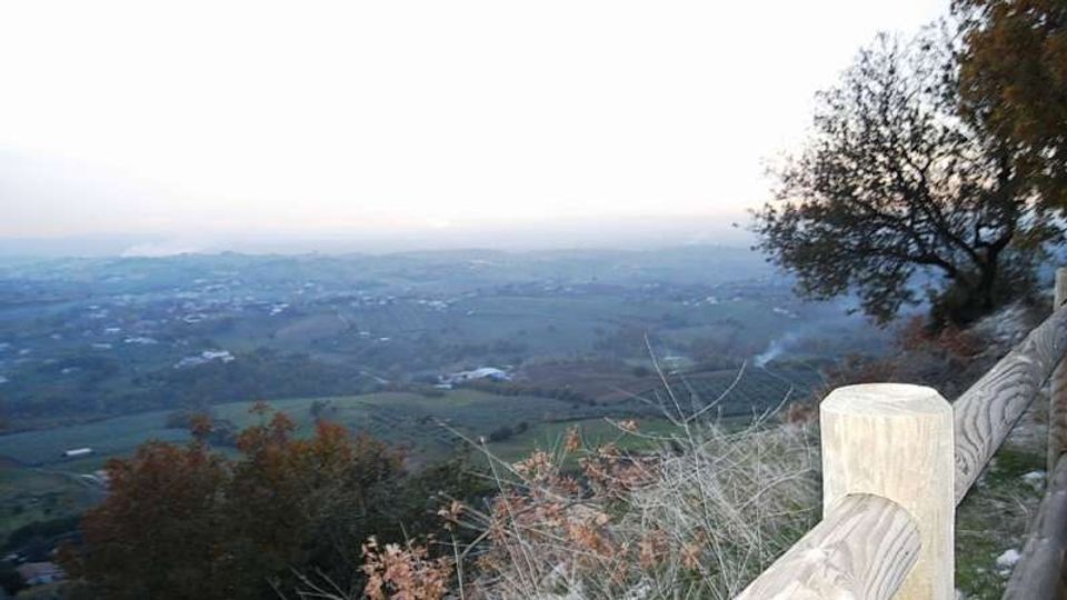 Belvedere su una collina con staccionata in legno in primo piano, affacciato su una vasta valle con campi e piccoli insediamenti. Vegetazione e alberi ai lati incorniciano l’orizzonte lontano sotto un cielo chiaro.